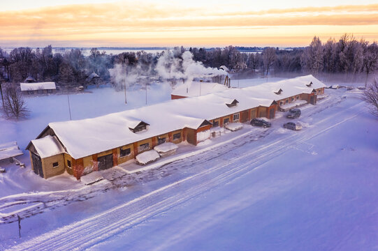 Aerial view of a long brick building covered in snow with smoke rising from a chimney, surrounded by a snowy landscape during golden hour in Latvija.