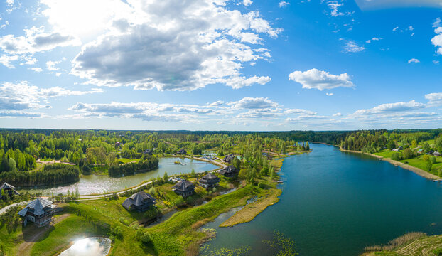 Aerial view of wooden houses nestled along a serene lake surrounded by lush green forests and rolling hills under a bright blue sky in Cesis, Cesis Municipality, Latvia.