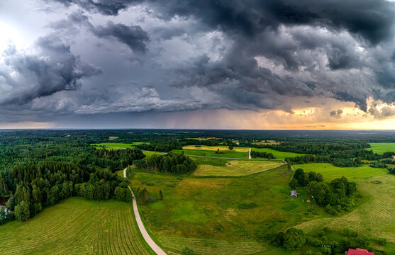 Aerial view of a rural landscape with lush green fields, dense forests, and a winding road under dramatic storm clouds during sunset in Latvia.