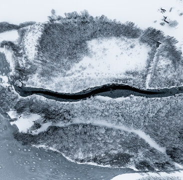 Aerial view of a winding river cutting through a snow-covered forest landscape in Latvia.