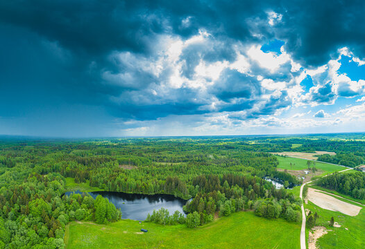 Aerial view of a serene lake nestled within a lush green forest and rolling meadows under a dramatic sky with heavy storm clouds in Latvia.