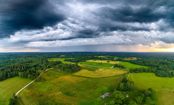 Aerial view of a rural landscape in Latvia featuring lush green fields, patches of forest, and a winding road under a dramatic dark storm sky with rain clouds in the distance, Latvia.