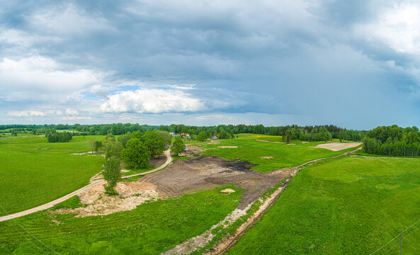 Aerial view of a rural landscape featuring lush green fields, a dirt path winding through trees, and a patch of dark soil under a dramatic cloudy sky in Latvia.