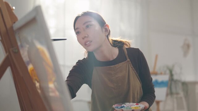Creative young Asian woman artist painting on canvas with brush in bright home studio. Relaxed female hobbyist enjoying peaceful indoor leisure time, expressing imagination for art.