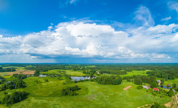 Aerial view of the lush green rural landscape featuring rolling fields, dense forests, small ponds, and a dramatic large cumulus cloud formation in the blue sky, Latvia.
