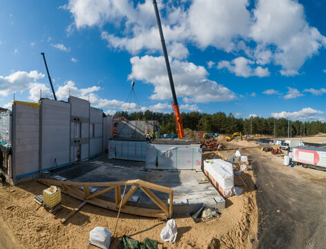 Aerial view of a construction site with a mobile crane lifting concrete panels, workers on site, and a forest background in Latvia.