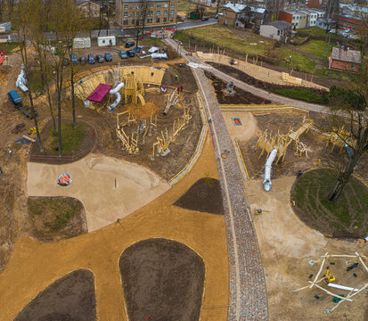 Aerial view of a wooden playground under construction with slides, sand pits, and walking paths surrounded by trees and residential buildings in Latvia.