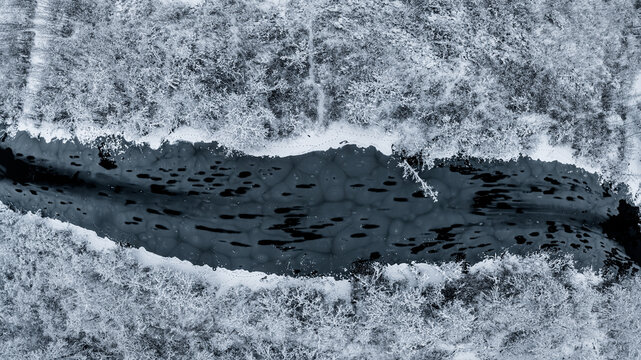 Aerial view of a frozen river with dark patterns on the ice surface, surrounded by a dense forest covered in fresh white snow in Latvia.