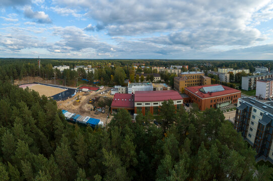 Aerial view of a school complex with a red roof, an adjacent sports field under construction, and surrounding pine forests in a residential area in Latvia.