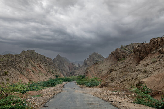 View of a narrow paved road winding through the rugged, rocky terrain of the Sulaiman Mountains under a dramatic, overcast sky in De-excluded Area Dera Ghazi Khan, Punjab, Pakistan.