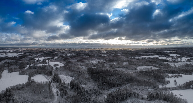 Aerial view of a snow-covered landscape featuring dense forests and open fields under a dramatic cloudy sky with sunlight breaking through, Latvia.