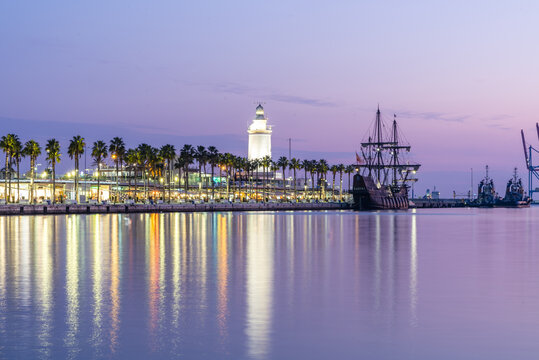 View of La Farola de Malaga lighthouse and a historic tall ship docked at the harbor at dusk with palm trees and city lights reflecting on the calm water in Malaga, Andalusia, Spain.