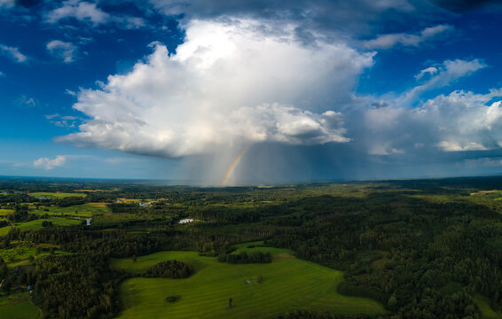 Aerial view of a vibrant rainbow arching over a lush green landscape with dense forests, rolling fields, and a dramatic storm cloud in Latvia.