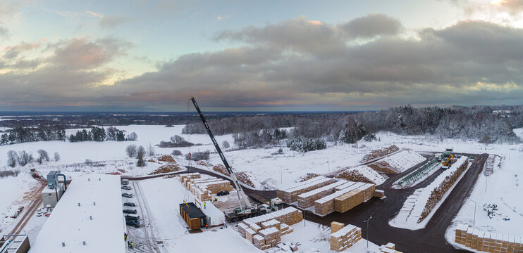 Aerial view of a lumber yard with stacks of timber and a mobile crane lifting a modular structure on a snow-covered industrial site in Latvia.