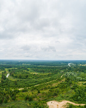 Aerial view of a lush green landscape with dense forests, a winding river, and dirt paths under a cloudy sky in Latvia.
