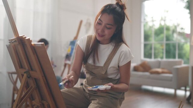 Creative young Asian woman artist painting on canvas with brush in bright home studio. Relaxed female hobbyist enjoying peaceful indoor leisure time, expressing imagination for art.