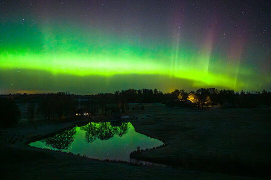 Aerial view of the aurora borealis illuminating the night sky with green and purple lights, reflecting over a calm pond surrounded by dark trees and a small building in Latvia.