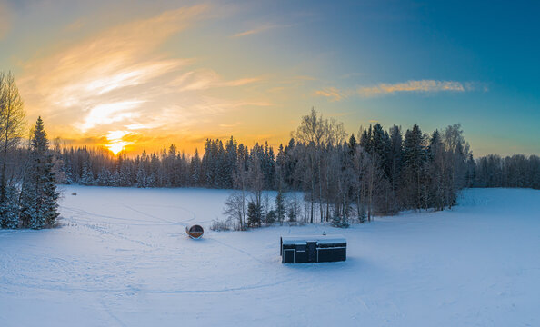 Aerial view of a snowy landscape featuring a small cabin and a barrel sauna nestled near a forest during a vibrant sunset in Latvia.