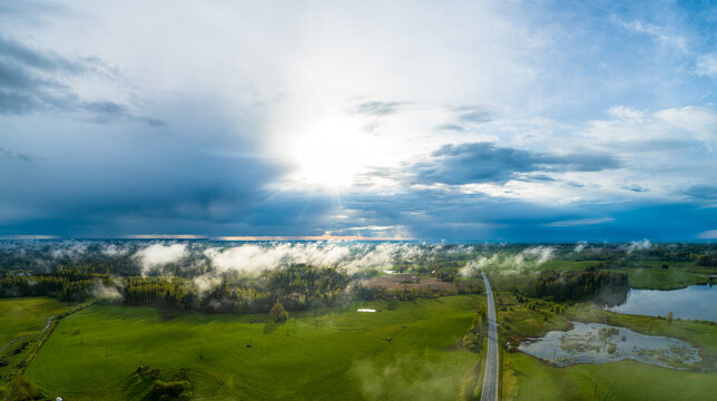 Aerial view of a rural landscape with green fields, a winding road, and a small lake under a dramatic sky with low-hanging clouds in Latvia.