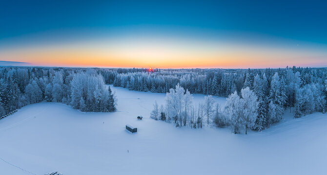 Aerial view of a snow-covered forest landscape with frost-covered trees and small wooden structures under a vibrant sunset sky in Latvia.