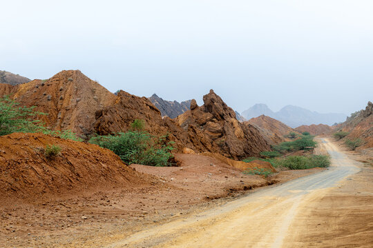 View of the rugged, reddish-brown rock formations of the Sulaiman Mountains, with a winding dirt road leading through the arid landscape in De-excluded Area Dera Ghazi Khan, Punjab, Pakistan.