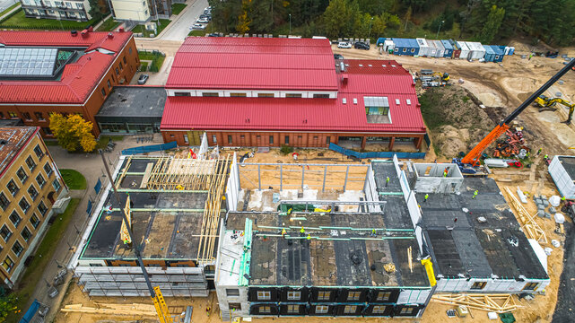 Aerial view of a construction site featuring a large building with a red roof, scaffolding, and workers active on the structure in Latvia.