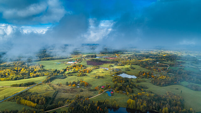 Aerial view of the rural landscape featuring lush green fields, patches of autumn-colored forests, small lakes, and dramatic clouds under a bright sky, Latvia.