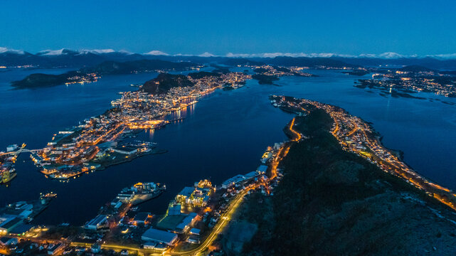 Aerial view of the illuminated city of Alesund at night, featuring coastal buildings, winding roads, and surrounding fjords in Alesund, More og Romsdal, Norway.