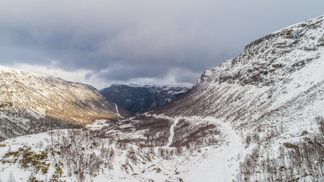 Aerial view of the snow-covered mountain valley near Flam, featuring winding roads, sparse trees, and dramatic rocky slopes in Flam, Vestland, Norway.