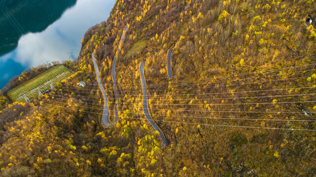 Aerial view of a winding mountain road surrounded by autumn foliage and dense forest near a fjord and an electrical substation in Flam, Vestland, Norway.