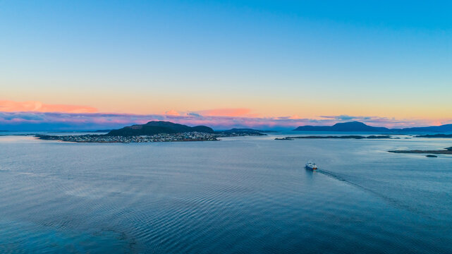 Aerial view of the coastal town of Alesund with a ferry sailing on the calm blue water under a sunset sky with pink clouds in Alesund, More og Romsdal, Norway.