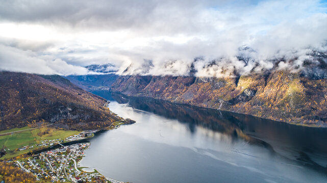 Aerial view of the village of Flam nestled along the Aurlandsfjord, surrounded by steep mountains with autumn foliage under a cloudy sky in Flam, Vestland, Norway.