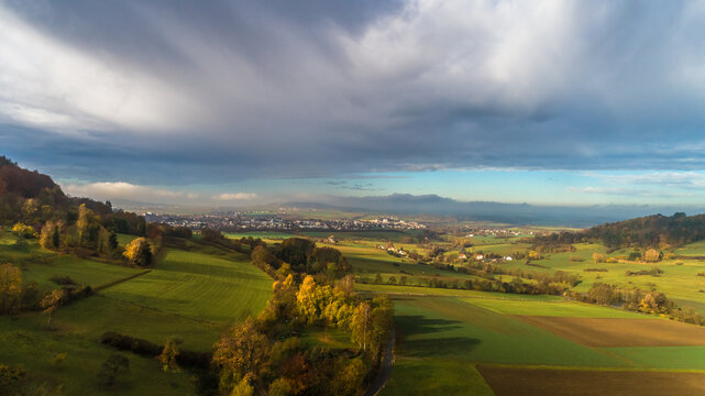 Aerial view of rolling green hills, agricultural fields, and patches of autumn trees under a dramatic cloudy sky in Germany.