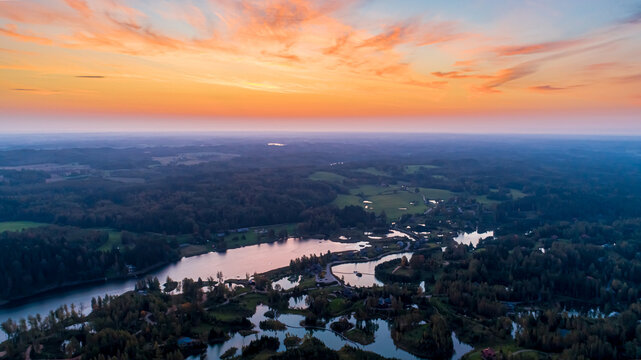 Aerial view of a serene lake surrounded by dense forests under a vibrant sunset sky in Latvia.