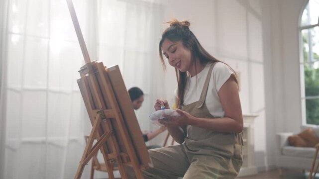 Creative young Asian woman artist painting on canvas with brush in bright home studio. Relaxed female hobbyist enjoying peaceful indoor leisure time, expressing imagination for art.