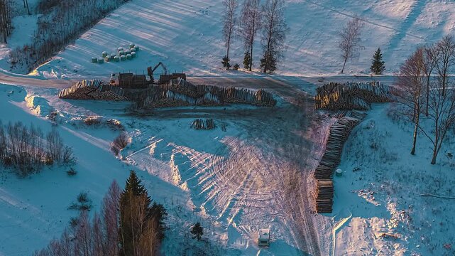 Aerial view of a logging site with stacked timber and machinery in a snowy landscape in Latvia.