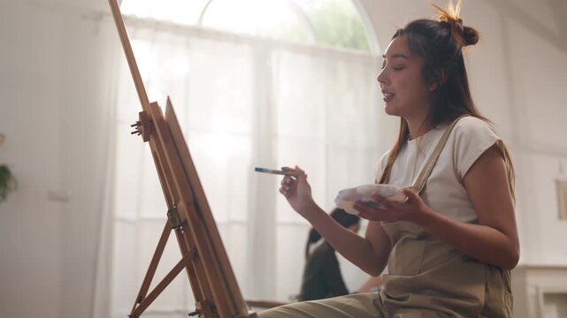 Creative young Asian women friends painting on canvas together in bright home studio. Relaxed female artists enjoying peaceful indoor leisure time, expressing imagination and passion for art.