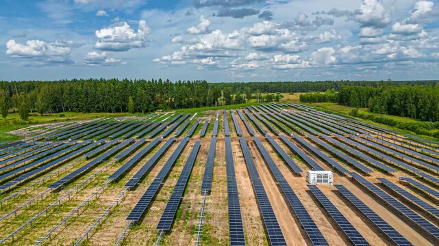 Aerial view of rows of solar panels installed in a rural field bordered by a dense pine forest under a bright blue sky with scattered clouds in Latvia.