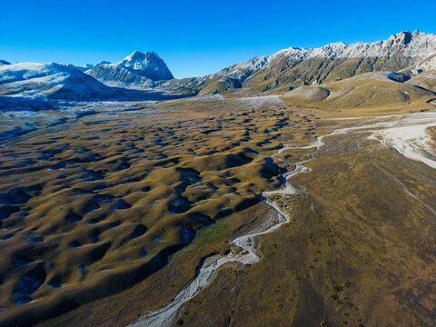 Aerial view of the undulating landscape of Gran Sasso d'Italia with a winding stream and snow-capped peaks under a clear blue sky in Pietracamela, Abruzzo, Italy.