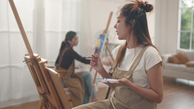 Creative young Asian women friends painting on canvas together in bright home studio. Relaxed female artists enjoying peaceful indoor leisure time, expressing imagination and passion for art.