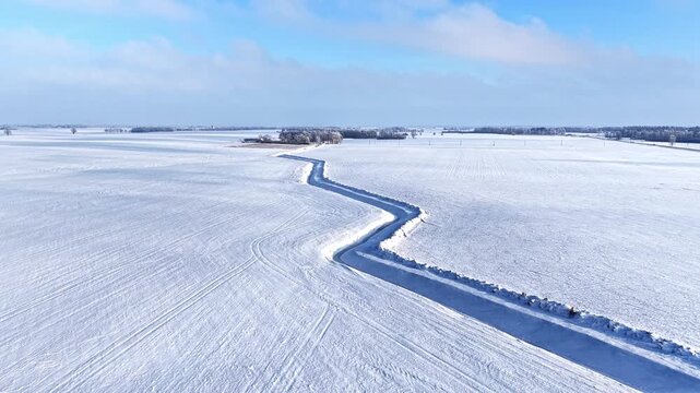 Aerial view of a winding road cutting through vast snow-covered fields under a clear blue sky in Latvia.