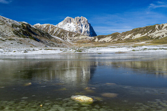 View of the frozen lake at the base of the snow-capped Gran Sasso d'Italia mountain under a clear blue sky in Pietracamela, Abruzzo, Italy.