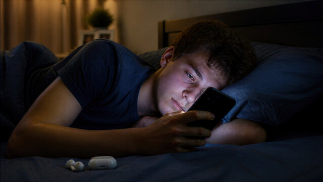 Young man lying in bed at night using smartphone in dark room with wireless earbuds