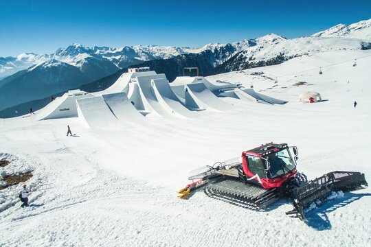 Aerial view of the Watles snow park with a red snow groomer on the slopes, featuring large snow jumps and surrounded by the Alps in Mals, Trentino-Alto Adige, Italy.
