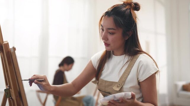 Creative young Asian women friends painting on canvas together in bright home studio. Relaxed female artists enjoying peaceful indoor leisure time, expressing imagination and passion for art.