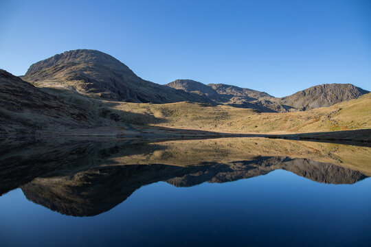 View of Styhead Tarn reflecting the surrounding mountains under a clear blue sky in Keswick, England, United Kingdom.