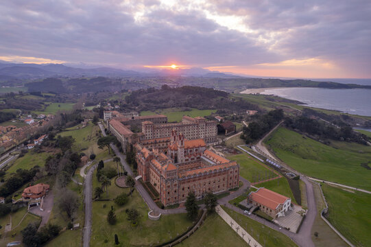 Aerial view of the historic Pontifical University of Comillas surrounded by lush green fields and the Cantabrian Sea at sunset in Comillas, Cantabria, Spain.