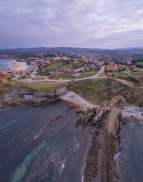 Aerial view of the coastal town of Comillas featuring rocky cliffs, a sandy beach, and historic architecture surrounded by green fields in Comillas, Cantabria, Spain.