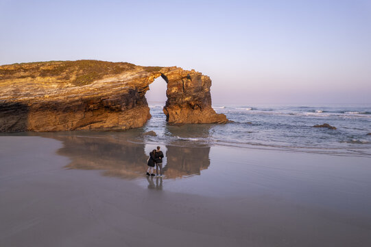 Aerial view of As Catedrais beach with a couple standing on the wet sand near the natural rock arch at golden hour in Spain.