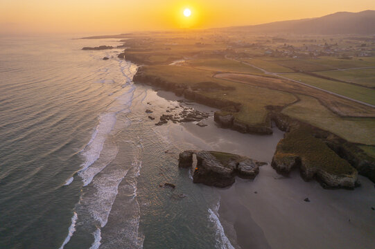 Aerial view of As Catedrais beach at sunset with golden light reflecting on the waves, rocky cliffs, and sandy coastline in Spain.